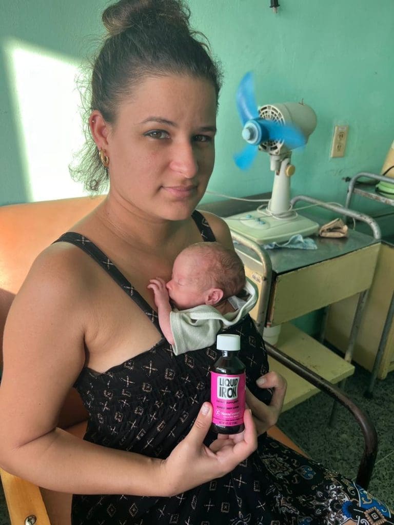 Mother in a black patterned dress sits in a clinic, breastfeeding her newborn who rests on her chest; she holds a pink-labeled bottle of Liquid Iron in her hand.