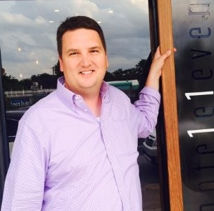 Man in a light purple button-down shirt standing at a glass door entrance, his hand resting on the door frame.