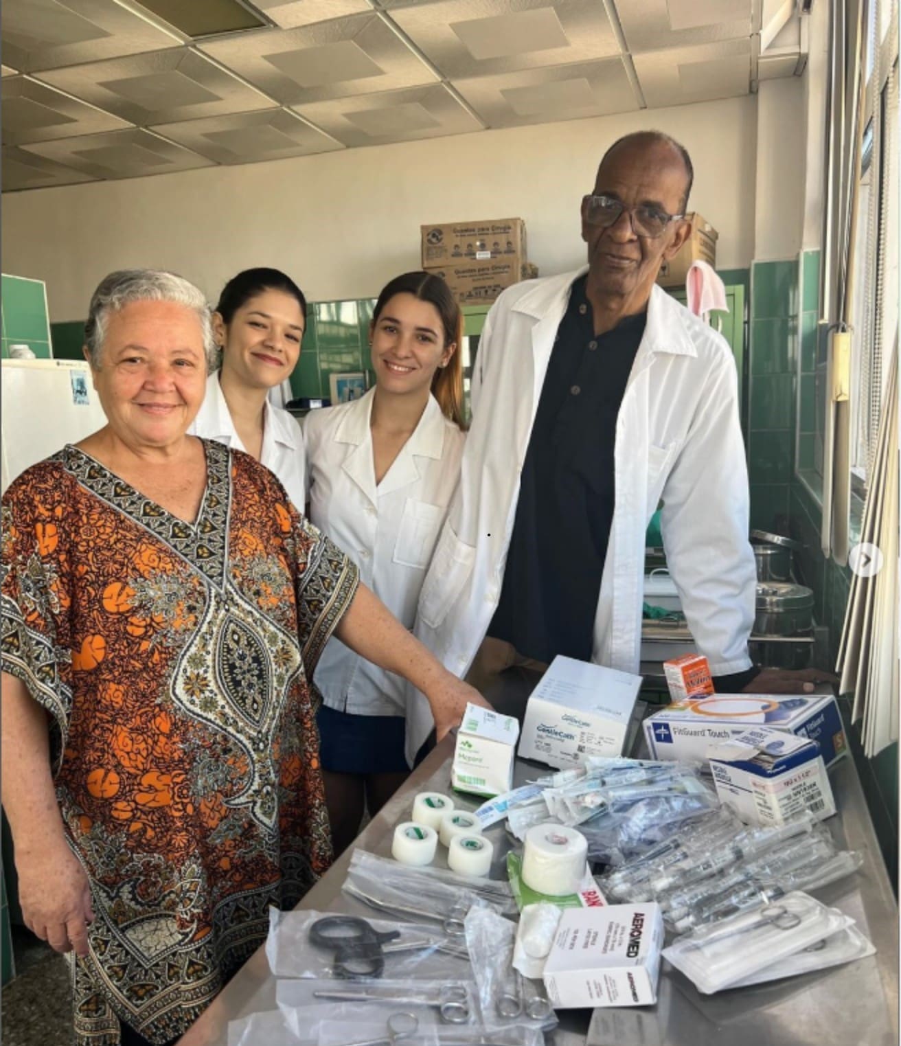 Smiling elderly woman in a patterned dress poses with three healthcare workers in white coats beside a table of medical supplies in a clinic setting.